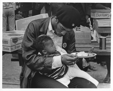 Black Panther Party member sits at a rally, feeding his baby son from his plate.